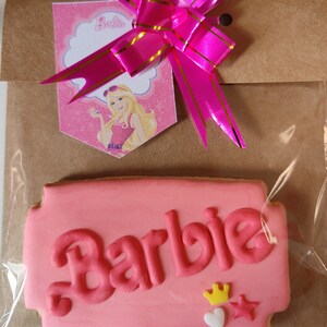 May include: Pink Barbie-themed cookie with the word "Barbie" in pink icing. The cookie is decorated with a yellow crown, a white heart, and a pink star.