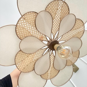 May include: A flower-shaped ceiling light fixture with a natural, bohemian design. The fixture features cream-colored fabric petals, woven rattan accents, and a central lightbulb. The light has a warm, inviting glow, perfect for adding a touch of nature-inspired elegance to any room.