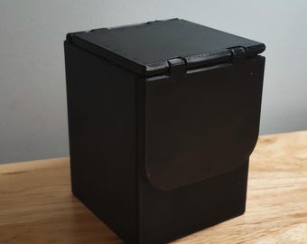 May include: A black, rectangular box with a hinged lid. The box is made of a solid material and sits on a wooden surface. The lid is closed, and the box appears to be empty. The box is a storage container.
