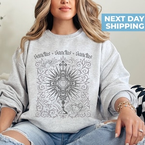 May include: A gray sweatshirt with a graphic print of a religious symbol, a chalice with a host, surrounded by a floral design. The text "Sanctus + Sanctus + Sanctus" is printed above the symbol.