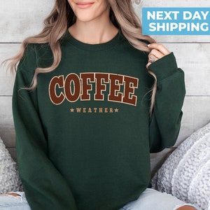 May include: A dark green sweatshirt with the text "COFFEE WEATHER" in brown letters. The sweatshirt is being worn by a person.
