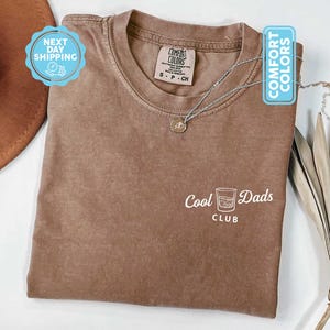 May include: Espresso-colored t-shirt with the text "Cool Dads Club" and a graphic of a cocktail glass. The shirt has a crew neck and short sleeves. A silver necklace with a pendant is draped across the shirt.