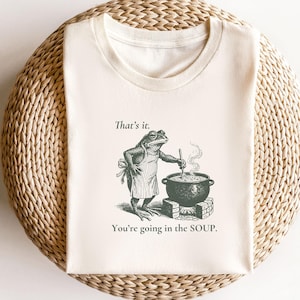 May include: A cream-colored t-shirt featuring a dark green graphic of a frog wearing an apron, stirring a large pot over a fire. The text above the frog reads "That's it." and the text below the pot reads "You're going in the SOUP."