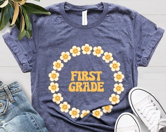 May include: A blue t-shirt with a white daisy flower wreath and the text "FIRST GRADE" in yellow letters.