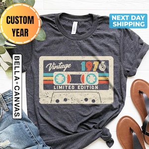 May include: Dark gray t-shirt featuring a vintage cassette tape design with the text "Vintage 1976 Limited Edition." The design includes retro colors and a distressed effect. The shirt is paired with blue jeans and brown sandals.