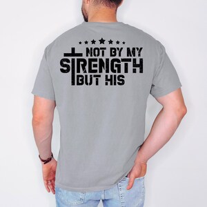 May include: A gray t-shirt with a black graphic that reads "Not by my strength but his" with a cross and stars.