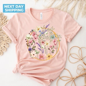 May include: A pink t-shirt with a floral design in a gold circle. The flowers are in shades of pink, yellow, blue, and purple.