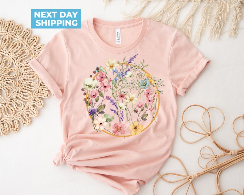 May include: A pink t-shirt with a floral design in a gold circle. The flowers are in shades of pink, yellow, blue, and purple.