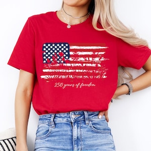May include: A red t-shirt featuring a distressed American flag design with white stars and stripes. The text "250 years of freedom" is printed below the flag. The shirt is made of a soft material.