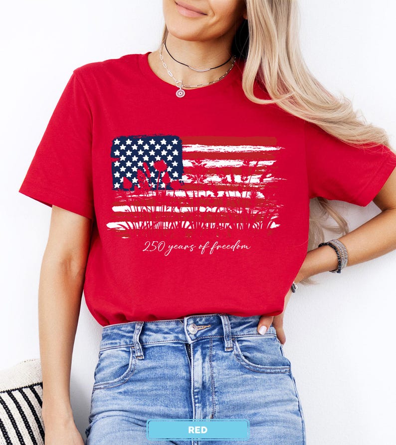 May include: A red t-shirt featuring a distressed American flag design with white stars and stripes. The text "250 years of freedom" is printed below the flag. The shirt is made of a soft material.