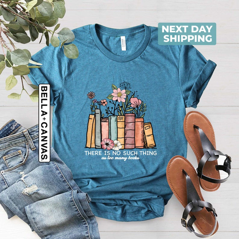 May include: Teal blue t-shirt with a graphic of a stack of colorful books with flowers growing out of them. The text "THERE IS NO SUCH THING as too many books" is printed below the graphic.