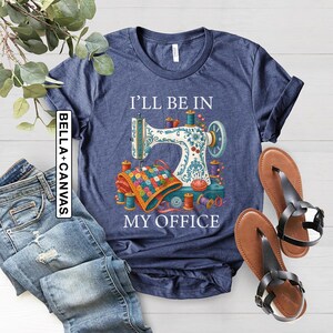 May include: Navy blue t-shirt with a sewing machine graphic and the text "I'll Be In My Office." The design includes spools of thread, a quilt, and a pincushion. A pair of brown sandals with black straps are next to the shirt.