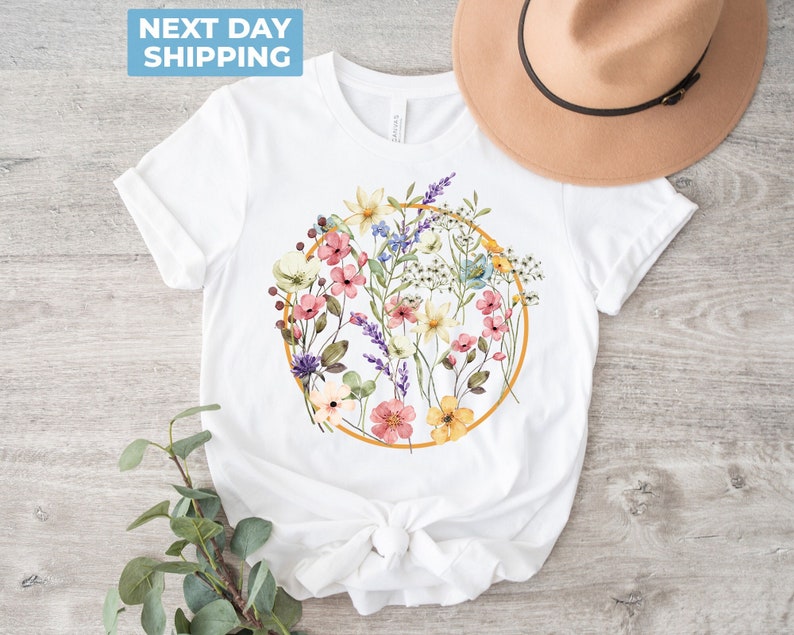 May include: White t-shirt with a circle design featuring a watercolor floral bouquet in shades of pink, yellow, blue, and green. The circle is outlined in gold.