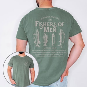 May include: A moss green t-shirt with the text "JESUS WILL MAKE YOU FISHERS OF MEN" and four fish illustrations. The text "MATTHEW 4:18-19" is below the fish illustrations. The shirt also has a small circular graphic on the front.