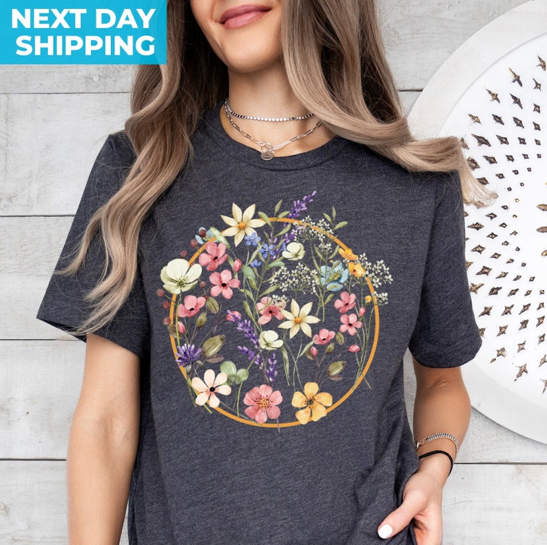 May include: A dark gray t-shirt with a floral design. The design features a circle of colorful flowers and greenery, surrounded by a gold outline.