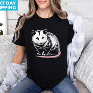 K&ouml;nnte beinhalten: Ein schwarzes T-Shirt mit einer wei&szlig;en und schwarzen Illustration eines Opossums. Das Opossum schaut den Betrachter an, mit leicht ge&ouml;ffnetem Maul. Das Opossum hat eine rosafarbene Nase und einen langen, rosafarbenen Schwanz.