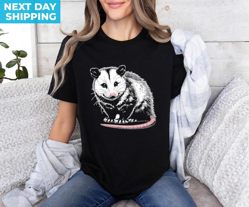 K&ouml;nnte beinhalten: Ein schwarzes T-Shirt mit einer wei&szlig;en und schwarzen Illustration eines Opossums. Das Opossum schaut den Betrachter an, mit leicht ge&ouml;ffnetem Maul. Das Opossum hat eine rosafarbene Nase und einen langen, rosafarbenen Schwanz.