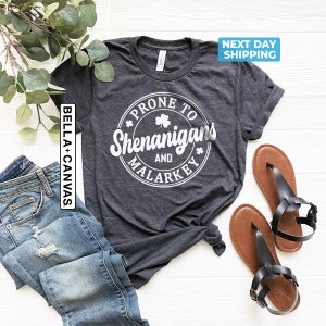May include: A dark grey t-shirt with a white circle graphic that says "Prone to Shenanigans and Malarkey" with a shamrock inside the circle.