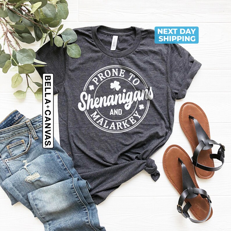 May include: A dark grey t-shirt with a white circle graphic that says "Prone to Shenanigans and Malarkey" with a shamrock inside the circle.