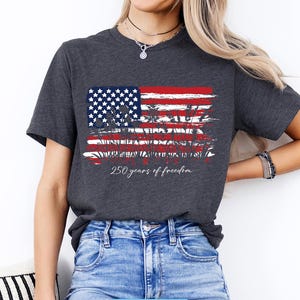 May include: Dark gray t-shirt featuring a distressed American flag design in red, white, and blue. The shirt includes the text "250 years of freedom" in a script font. The shirt is a crew neck style.