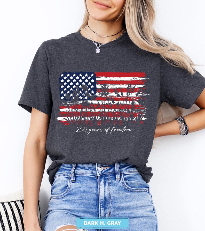 May include: Dark gray t-shirt featuring a distressed American flag design in red, white, and blue. The shirt includes the text "250 years of freedom" in a script font. The shirt is a crew neck style.