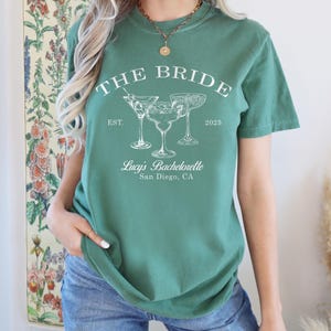 May include: A green t-shirt with the text "The Bride Est. 2025" and three cocktail glasses. The text "Lucy's Bachelorette San Diego, CA" is also on the shirt.