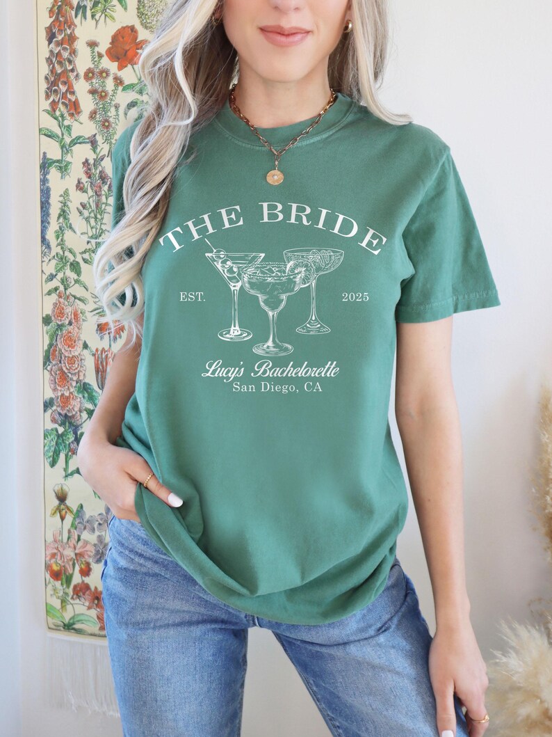 May include: A green t-shirt with the text "The Bride Est. 2025" and three cocktail glasses. The text "Lucy's Bachelorette San Diego, CA" is also on the shirt.