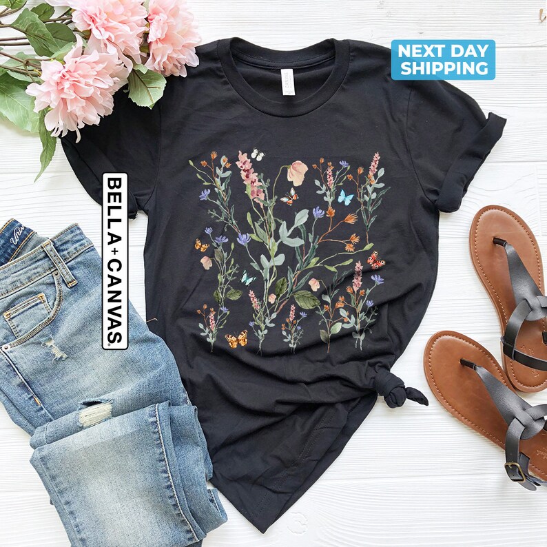 May include: Black t-shirt with a floral print featuring a variety of flowers and butterflies in shades of pink, blue, green, and orange. The text "Bella + Canvas" is printed on the left side of the shirt.