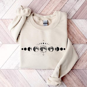 May include: A cream-colored sweatshirt with a black graphic of a moon phases design. The design features a crescent moon, half moon, full moon, and other phases of the moon.