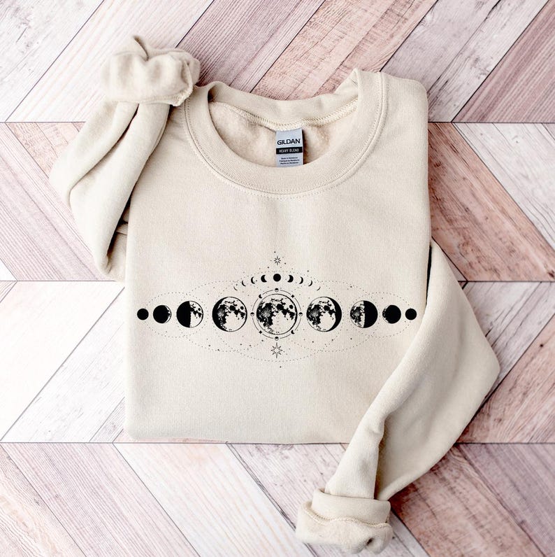 May include: A cream-colored sweatshirt with a black graphic of a moon phases design. The design features a crescent moon, half moon, full moon, and other phases of the moon.