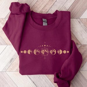 May include: Burgundy sweatshirt with a gold moon phase design. The design features a crescent moon, full moon, and other phases of the moon.