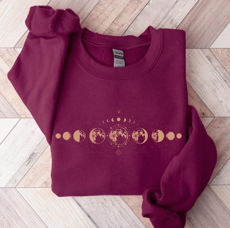 May include: Burgundy sweatshirt with a gold moon phase design. The design features a crescent moon, full moon, and other phases of the moon.
