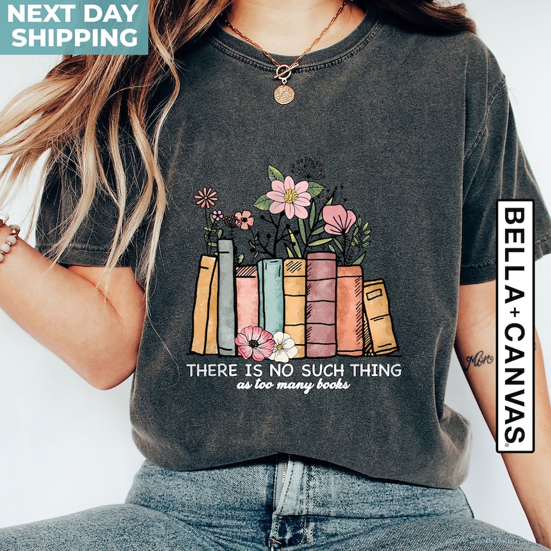 May include: A dark gray t-shirt with a graphic of a stack of colorful books with flowers on top. The text "THERE IS NO SUCH THING as too many books" is printed below the graphic.