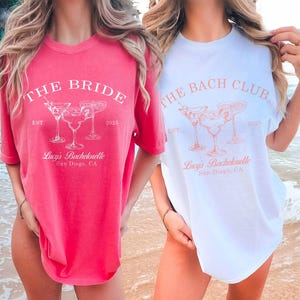 May include: Two oversized t-shirts with a bachelorette party theme. The pink shirt says "The Bride" with a margarita graphic and the white shirt says "The Bach Club" with a margarita graphic. Both shirts say "Lucy's Bachelorette" and "San Diego, CA".