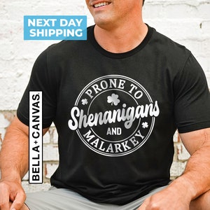 May include: Black t-shirt with a silver circle graphic that says "Prone to Shenanigans and Malarkey" with a shamrock design.