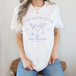 May include: White t-shirt with blue text that reads "The Bach Club Est. 2025" and a graphic of three martini glasses. The text below the graphic reads "Lucy's Bachelorette San Diego, CA."