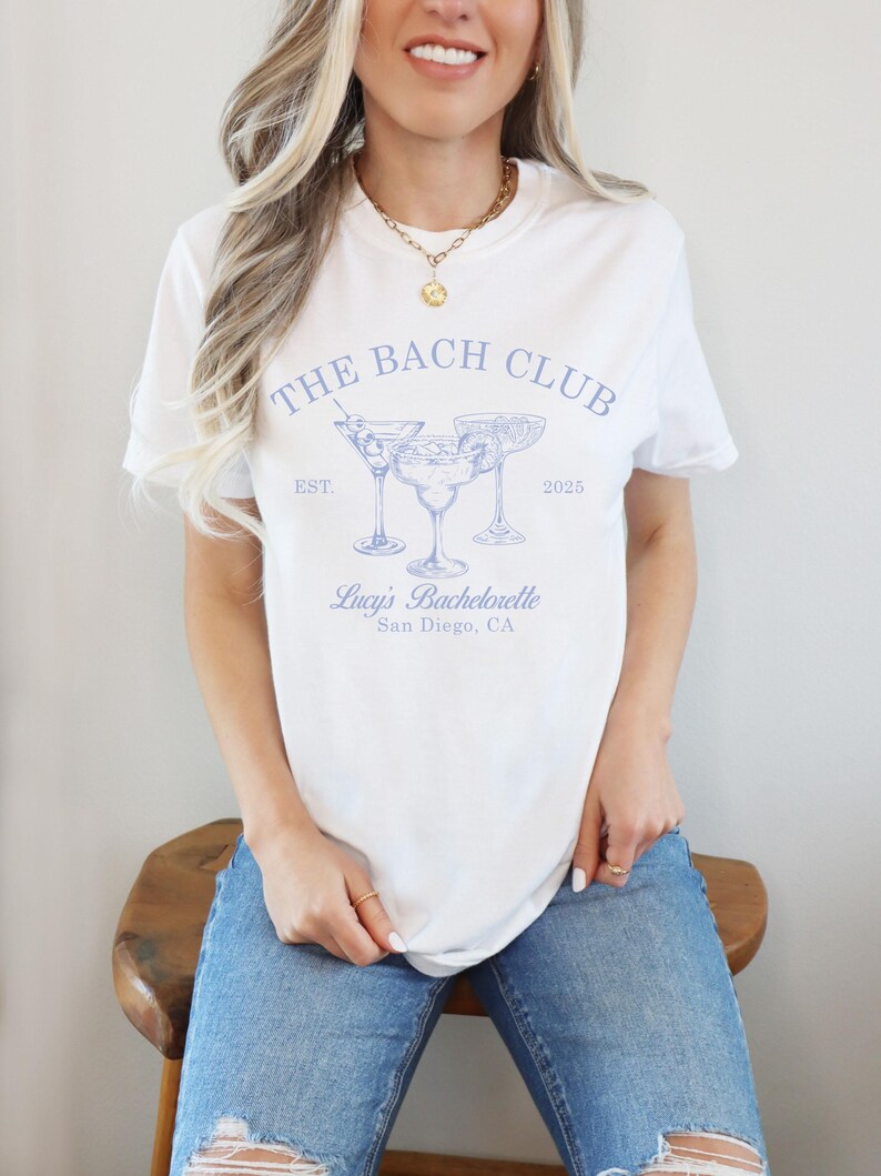 May include: White t-shirt with blue text that reads "The Bach Club Est. 2025" and a graphic of three martini glasses. The text below the graphic reads "Lucy's Bachelorette San Diego, CA."