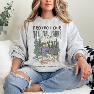 May include: A gray sweatshirt with the text "PROTECT OUR NATIONAL PARKS" above a graphic of a mountain scene and a fish. The sweatshirt also has the text "EXPLORE RESPONSIBLY". The person is wearing ripped blue jeans.