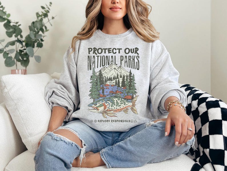 May include: A gray sweatshirt with the text "PROTECT OUR NATIONAL PARKS" above a graphic of a mountain scene and a fish. The sweatshirt also has the text "EXPLORE RESPONSIBLY". The person is wearing ripped blue jeans.