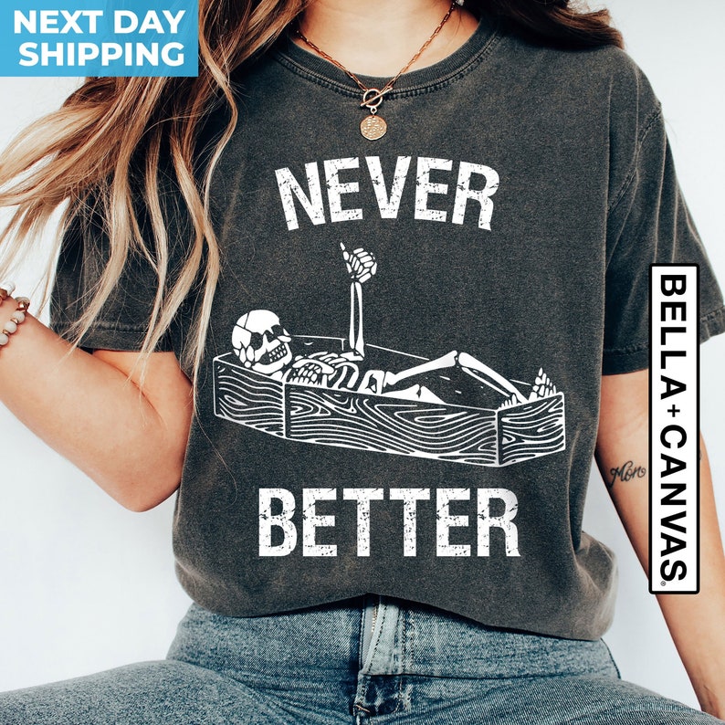 May include: A dark gray t-shirt with a white graphic of a skeleton lying in a coffin with the text "NEVER BETTER" above it.