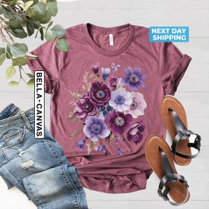 May include: A maroon t-shirt featuring a floral design with purple, lavender, and white anemone flowers. The shirt is paired with blue jeans and brown sandals. The text "BELLA + CANVAS" is on the left side of the shirt.
