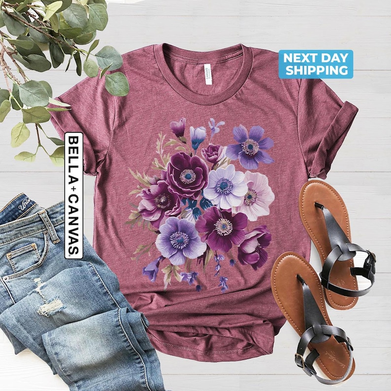 May include: A maroon t-shirt featuring a floral design with purple, lavender, and white anemone flowers. The shirt is paired with blue jeans and brown sandals. The text "BELLA + CANVAS" is on the left side of the shirt.