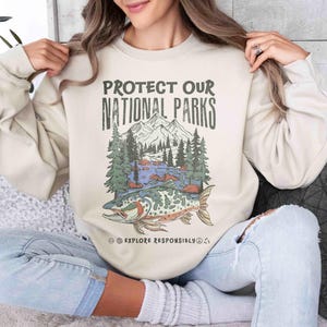 May include: Beige sweatshirt with the text "PROTECT OUR NATIONAL PARKS" above a graphic of a mountain scene with a fish. The graphic includes trees, mountains, and a river. The words "EXPLORE RESPONSIBLY" are below the graphic.
