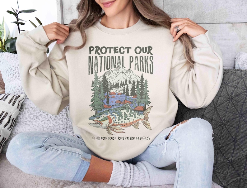 May include: Beige sweatshirt with the text "PROTECT OUR NATIONAL PARKS" above a graphic of a mountain scene with a fish. The graphic includes trees, mountains, and a river. The words "EXPLORE RESPONSIBLY" are below the graphic.