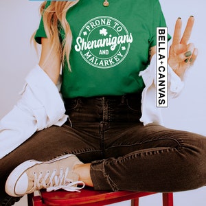 May include: A green t-shirt with a white circle graphic that says "Prone to Shenanigans and Malarkey" with a shamrock design. The shirt is being modelled by a person wearing black jeans and white trainers.