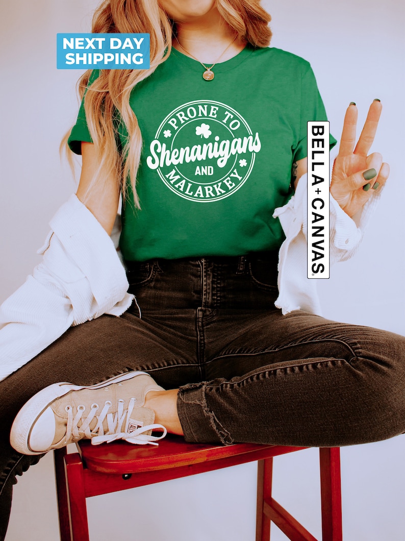 May include: A green t-shirt with a white circle graphic that says "Prone to Shenanigans and Malarkey" with a shamrock design. The shirt is being modelled by a person wearing black jeans and white trainers.
