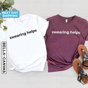 Op de afbeelding: Twee T-shirts met de tekst "swearing helps" erop gedrukt. E&eacute;n shirt is wit en de andere is bordeaux.