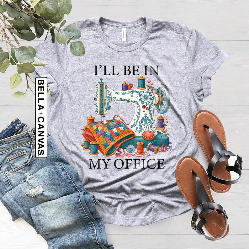 May include: A light gray Bella+Canvas t-shirt featuring a colorful sewing machine graphic with the text "I'll Be In My Office." The graphic includes spools of thread, a quilt, and floral accents. The shirt is paired with blue jeans and brown sandals.