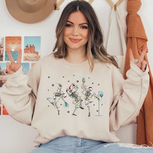 May include: A beige sweatshirt with a festive design featuring three skeletons dressed in Christmas lights and hats. The skeletons are holding drinks and a IV bag.