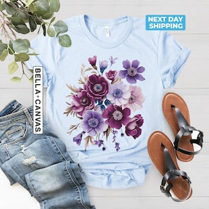 May include: Light blue t-shirt with a floral design in shades of purple and white. The shirt has the text "BELLA + CANVAS" on the side. A pair of brown sandals with black straps are next to the shirt.